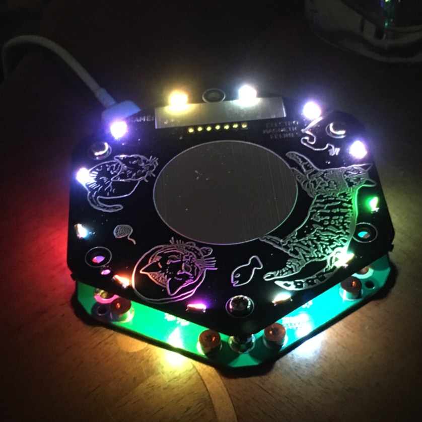 A hexagonal camp badge, made up of two PCBs with a gap between them, lit by RGB LEDs on top, with more LEDs shining inside. It has illustrations of cats on the silkscreen.