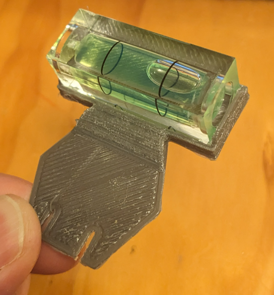 A small spirit level, attached to a 3D printed hexagonal mount with notches to attach it to the badge. The mounting hexagon is slightly larger than the thumb holding it.